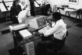 图中是一个办公室场景，包括一位站在早期个人电脑前的员工
