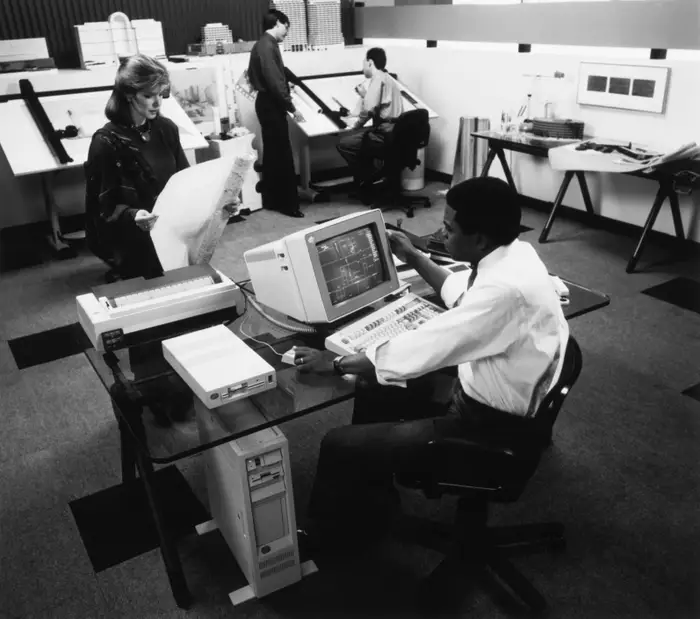图中是一个办公室场景，包括一位站在早期个人电脑前的员工